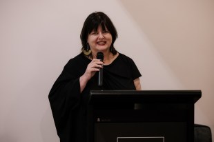 woman with dark hair in black dress, smiling and giving speech in gallery. ADC