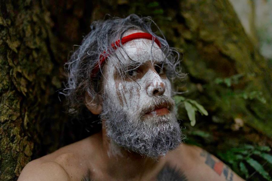 Jake Māra in a promotional image for ‘Arawarra’, part of Merrigong Theatre Company's MERRIGONGX 2026 program. A bearded Aboriginal man, his face painted with white ochre and wearing a red headband, looks steadily at the camera.