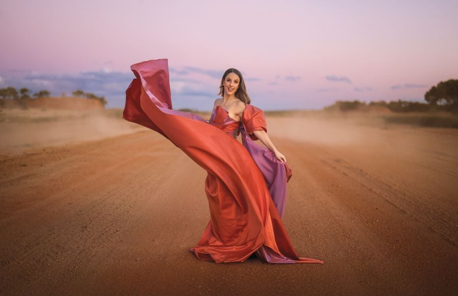 Promotional image for Opera Queensland's Festival of Outback Opera. A fair-skinned woman stands in the middle of a wide dirt road at dusk, her billowing theatrical costume in stark contrast with the regional setting.
