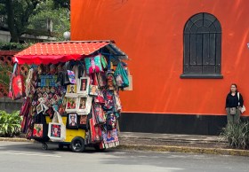 A vendor's cart on street filled with Frida Kahlo souvenirs with red building behind.