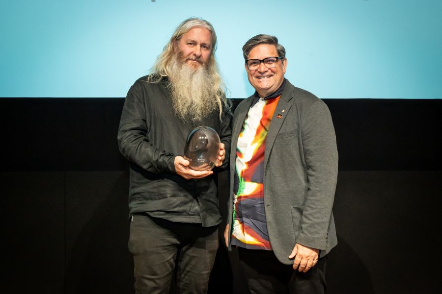Robin Fox, the recipient of the Creative Australia Award for Emerging Experimental Arts, and Wesley Enoch AM, Chair of the Australia Council Board of Creative Australia, at the Creative Australia Awards 2026 ceremony. A tall, fair-skinned man with long, greying fair hair and a long grey beard, holds a large glass award in his hands. To his left stands an middle-aged Aboriginal man with greying black hair and wearing black glasses, a colourful t-shirt and a charocal-coloured jacket over a colourful t-shirt. 