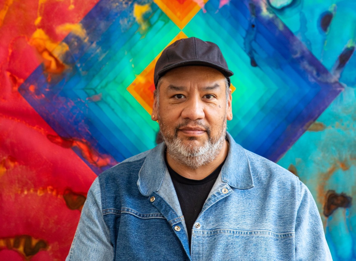 Jeffrey Gibson. Indigenous man in denim shirt and black cap in front of colorful painting.