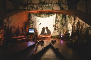 A scene from Australian Theatre for Young People's 'The Last Train to Madeline'. Two young people silhouetted behind a hanging sheet, one female, one male, lean towards each other as if for a kiss. They sit on a stage dressed with artificial plants, a small television and other objects.