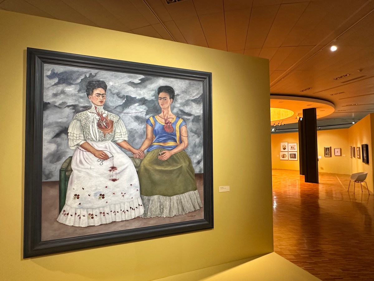 yellow wall of gallery showing painting of two women sitting on chairs holding hands in Surreal style. Frida Kahlo