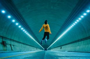 A parkour practioner dressed in basecall cap, yellow hoodie, black pants and white sneakers, leaps into the air in a tunnel which appears to be closed to traffic. Blue-white lights in a row along the tunnels walls receed into the distance, with the leaping body centred in the frame. the photo illustrates ArtsHub's weekly round-up of arts sector appointments.