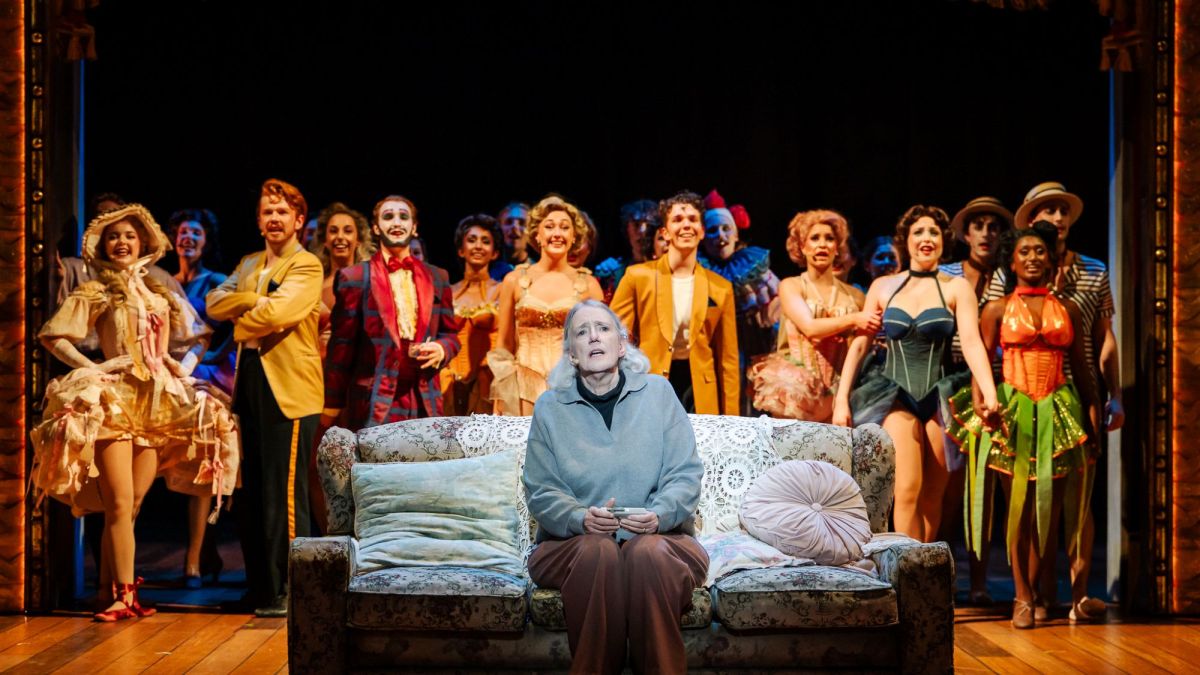 A scene from WAAPA's new Australian musical by Eddie Perfect, 'Tivoli Lovely'. An elderly, grey-haired woman in a grey cardigan sits on a sofa in a nursing home as her showgirl memories of life on the Tivoli Circuit come to colourful life behind her.