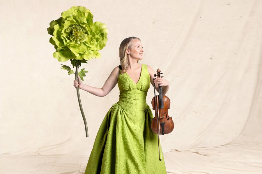 Skye McIntosh, the Australian Haydn Ensemble's Artistic Director, Founder and Principal Violinist. A fair-skinned woman with long blonde hair poses in profile. She wears a green gown, holds a violin in her left hand and a giant green carnation in her right hand.