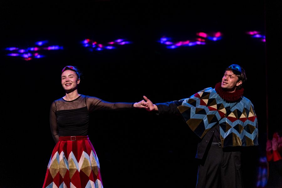 A scene from Bell Shakespeare's 2025 production of 'Romeo and Juliet'. Two young people hold hands at a masquerade ball, their masks raised to show their smiling faces.