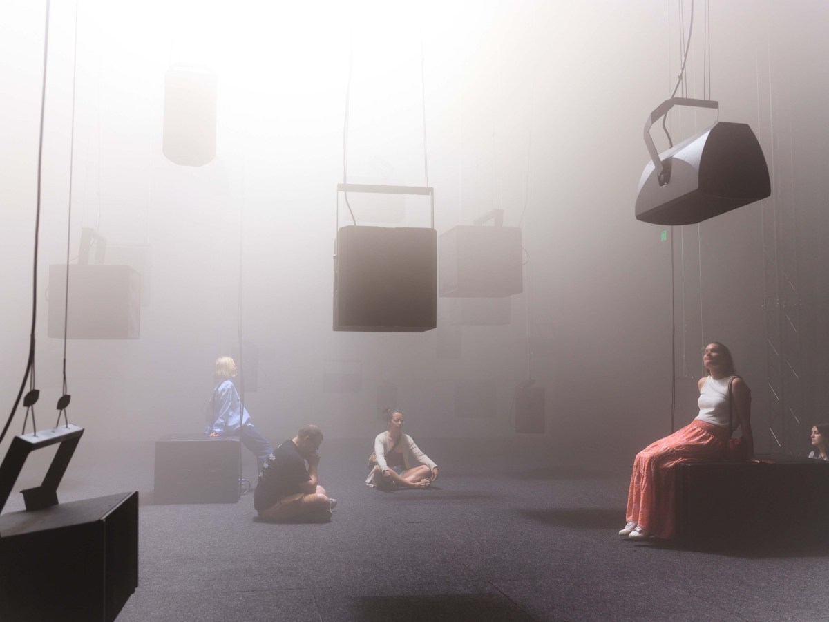 Fremantle Biennale 2025: a large smokey empty room with 7 large black square shaped speakers suspended from the room while three young people are sitting on the floor looking at them.