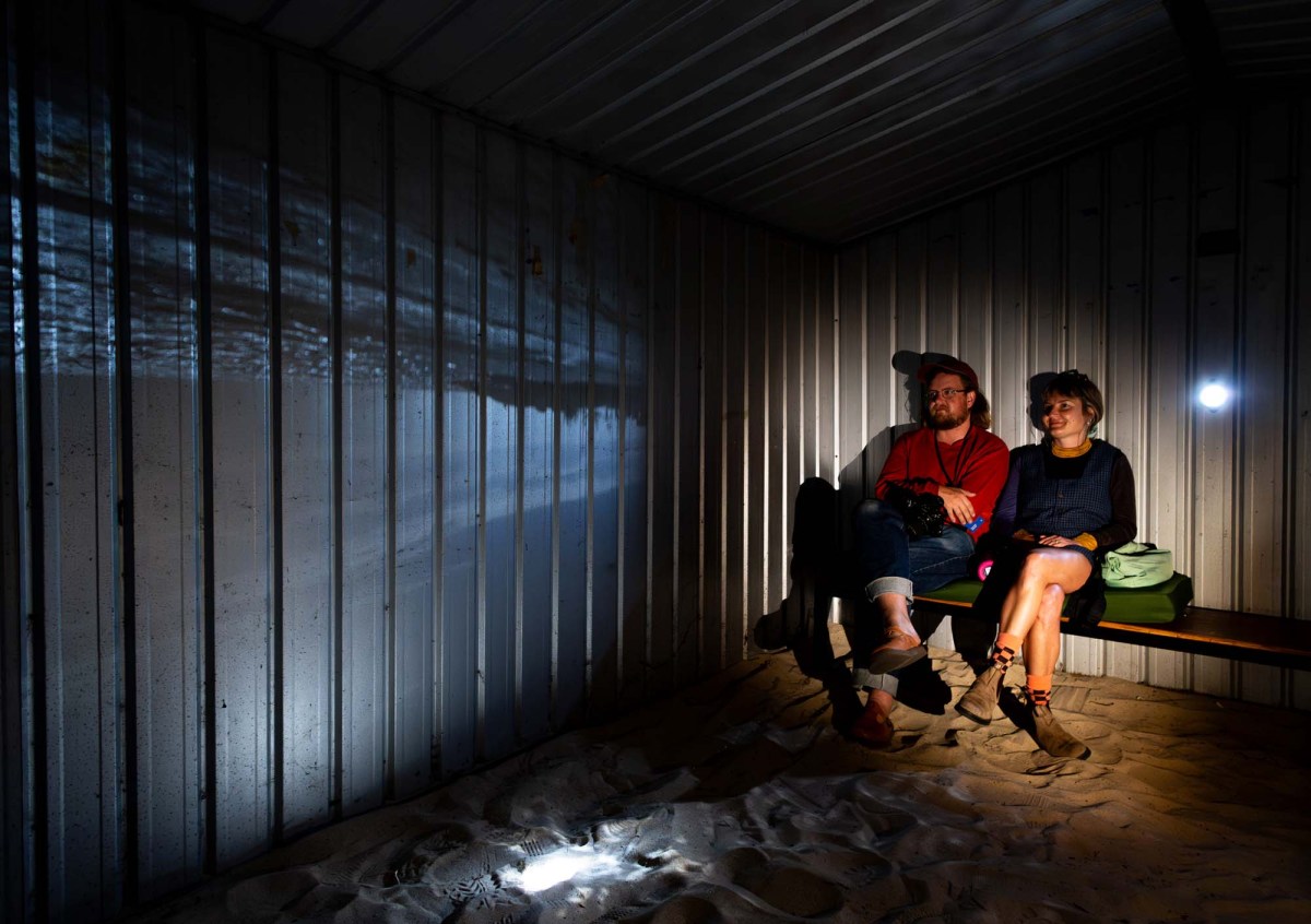 Fremantle Biennale: A young man and woman sitting inside on a bench in a small tin shed in the dark looking at a projected image on one of its inside walls.