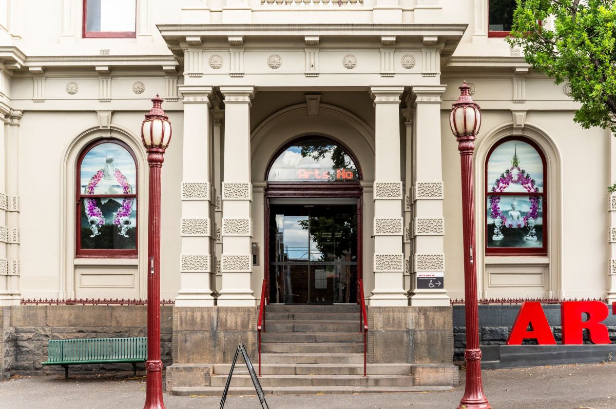 The front door of Arts House, North Melbourne Town Hall, with Nathan Beard's 'Floral Histories' installed. The portico of a Victorian-era building flanked by two old-fashioned lamp posts; floral artworks are visible in the windows on either side of the portico.