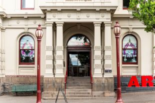 The front door of Arts House, North Melbourne Town Hall, with Nathan Beard's 'Floral Histories' installed. The portico of a Victorian-era building flanked by two old-fashioned lamp posts; floral artworks are visible in the windows on either side of the portico.