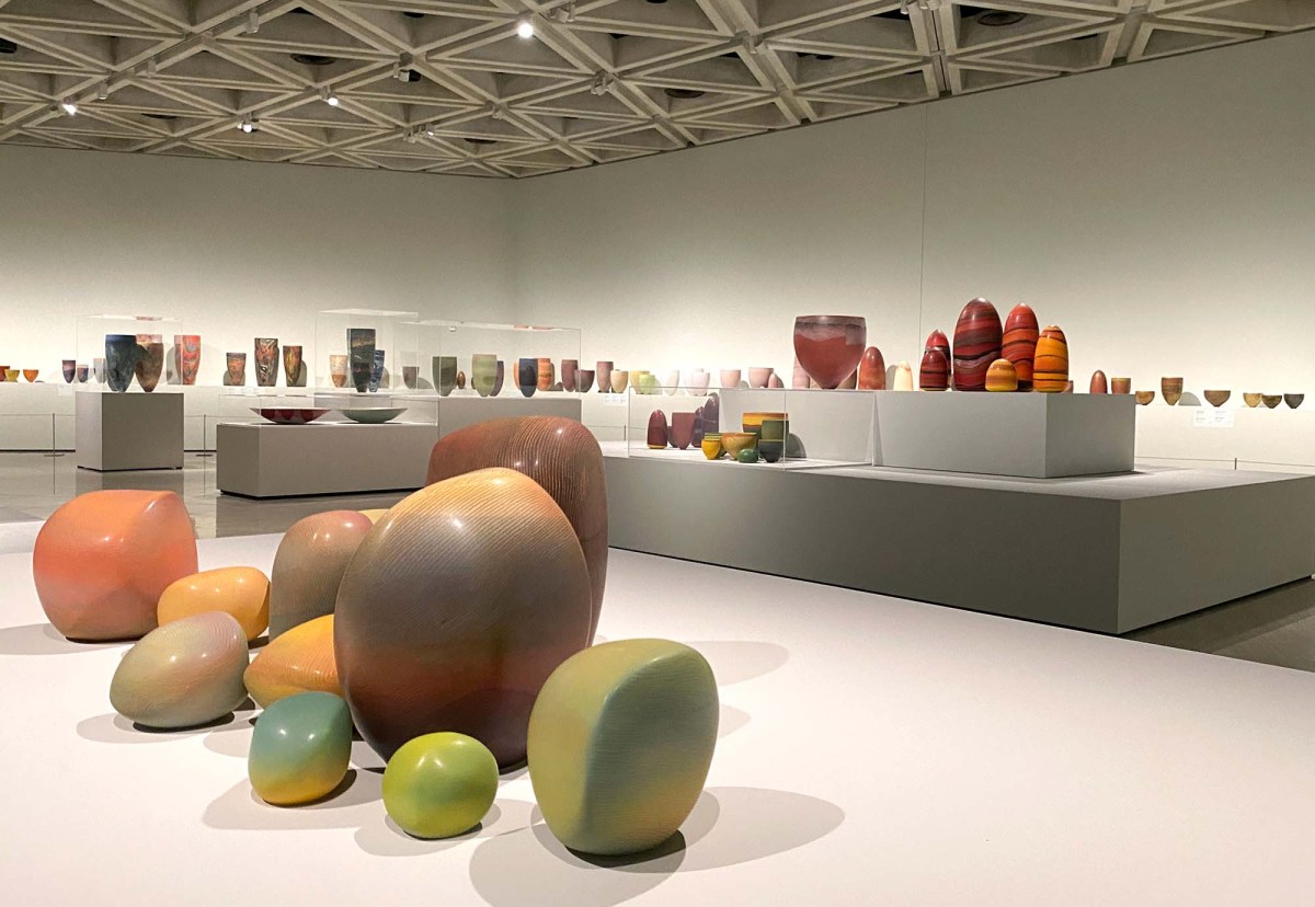 Pippin Drysdale Infinite Terrain: a photo of a ceramics exhibition in a large contemporary gallery space. The ceramics are colourful vessels and stone-like shapes of various sizes displayed on low to the ground wide shelf-like blocks and medium sized narrower plinths.