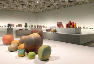 Pippin Drysdale Infinite Terrain: a photo of a ceramics exhibition in a large contemporary gallery space. The ceramics are colourful vessels and stone-like shapes of various sizes displayed on low to the ground wide shelf-like blocks and medium sized narrower plinths.