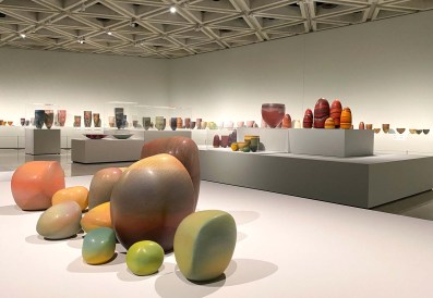 Pippin Drysdale Infinite Terrain: a photo of a ceramics exhibition in a large contemporary gallery space. The ceramics are colourful vessels and stone-like shapes of various sizes displayed on low to the ground wide shelf-like blocks and medium sized narrower plinths.