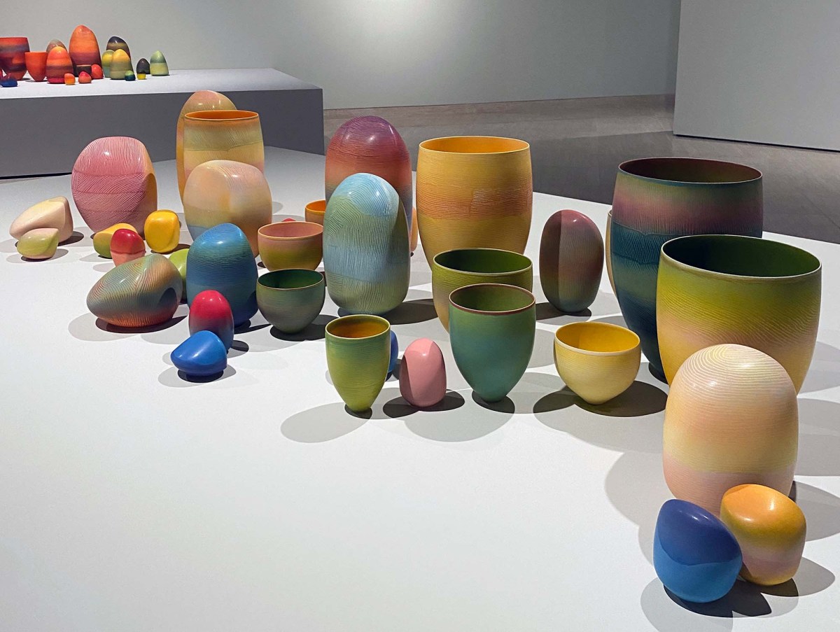 Pippin Drysdale: Infinite Terrain Major Retrospective exhibition 2025: a photo of brightly coloured ceramic vessels and stone like forms in shades of light blue, yellow, pink and green displayed on a low to the ground, wide platform plinth in a contemporary gallery space.