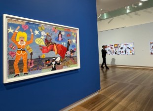 view of gallery with blue wall and bright coloured painting in comic style. Kaylene Whiskey