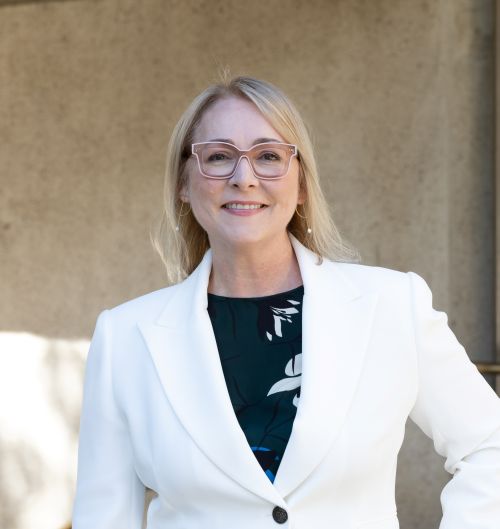 Renai Grace is the new CEO of Queensland Museum. A fair-haired, fair-skinned woman wearing glasses and a white jacket over a black shirt decorated with bold geometric designs.