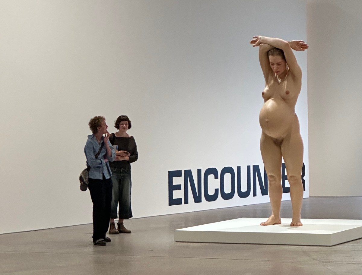 Two women looking at hyperreal sculpture of pregnant woman in gallery setting. Ron Mueck