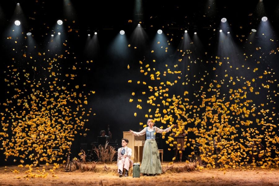 Cameron Bajraktarevic-Hayward and Kala Gare in MTC's 2026 production of My Brilliant Career. Photo: Pia Johnson. A rain of flowers falls onto a stage covered by dry grass; Gare as Sybylla spreads her arms and mouth in amazement while Bajraktarevic-Hayward sits primly beside her.