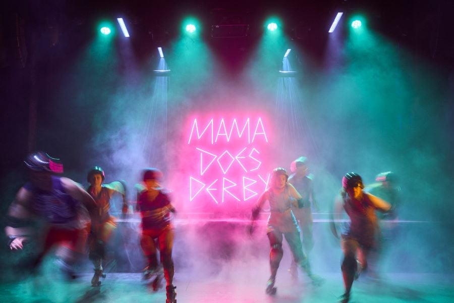 Mama Does Derby features actual roller derby players alongside professional actors. The photo depicts blurred skaters emerging at speed from theatrical smoke; a bright pink neon sign behind them reads 'MAMA DOES DERBY' in capital leads.