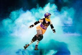 A woman in a bright roller derby uniform and protective gear, including pads and helmets, skates out of a cloud of theatrical smoke. Mama Does Derby