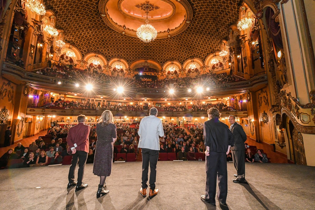 Sydney Film Festival is accepting applications for 2026.  The photo depicts a special event at the festival, with five guests standing on the stage of the State Theatre looking out at a crowded and brightly lit auditorium. opportunities awards funding