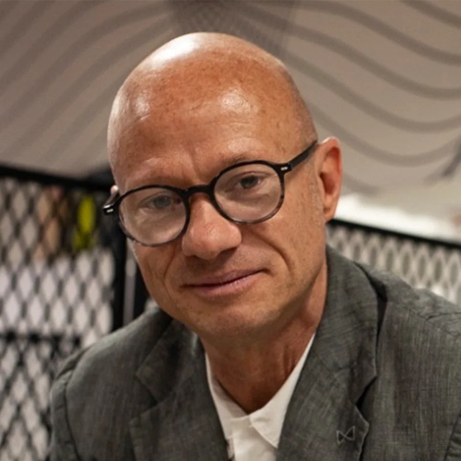 Noel Jordan joins Monkey Baa Theatre Company as General Manager. Noel  is an older man with a shaved head; he wears dark framed glasses, a grey linen jacket and a white shirt. 