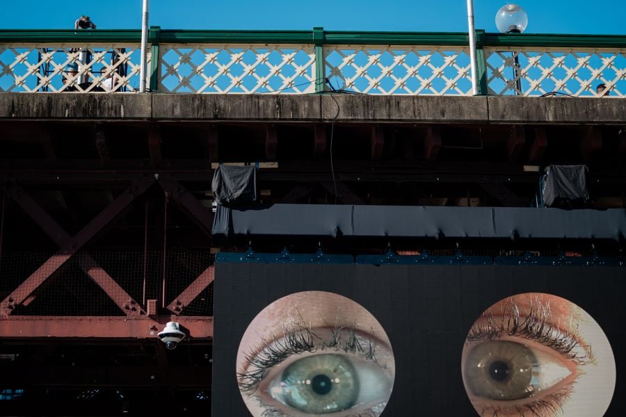 Julia Phillips’ Observer, Observed at Sydney Festival 2026. Installation view of the artwork: at top right, a person peers down from atop the bridge through binoculars at the photographer; their eyes, picked up by cameras inside the binoculars, are projected larger than life on an LED screen beneath the bridge.