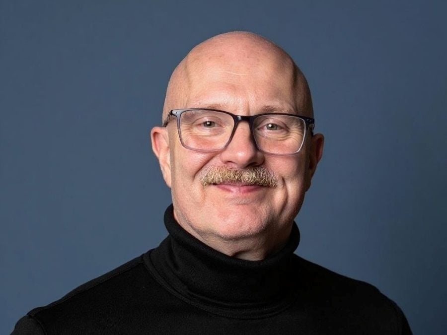 Darwin Symphony Orchestra's new General Manager, Paul Christ. A fair-skinned, older man with a shaved head, glasses, and a small, neat moustache. He wears a black turtleneck jumper and is smiling at the camera.