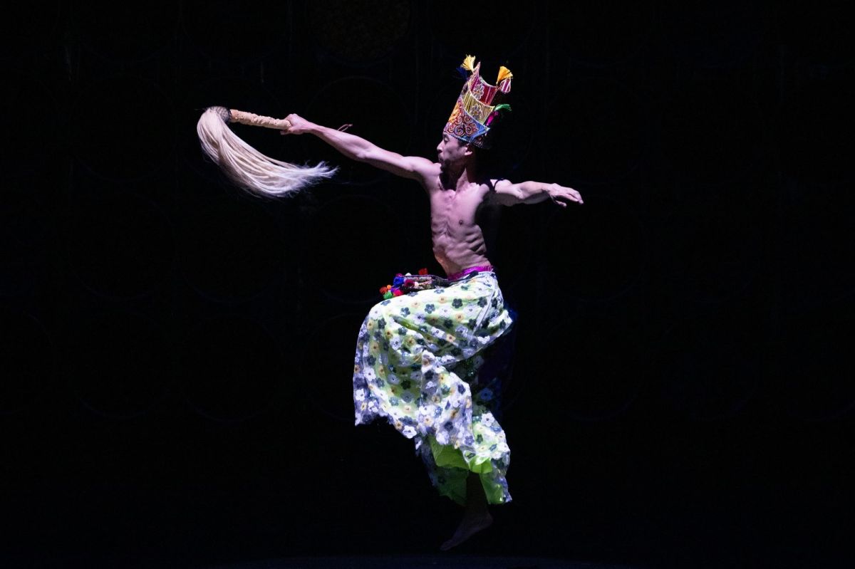 A dance sequence from Post-Orientalist Express, 2026. A shirtless male dancer poised on one foot with his arms extended; he wears a colourful headdress and costume.