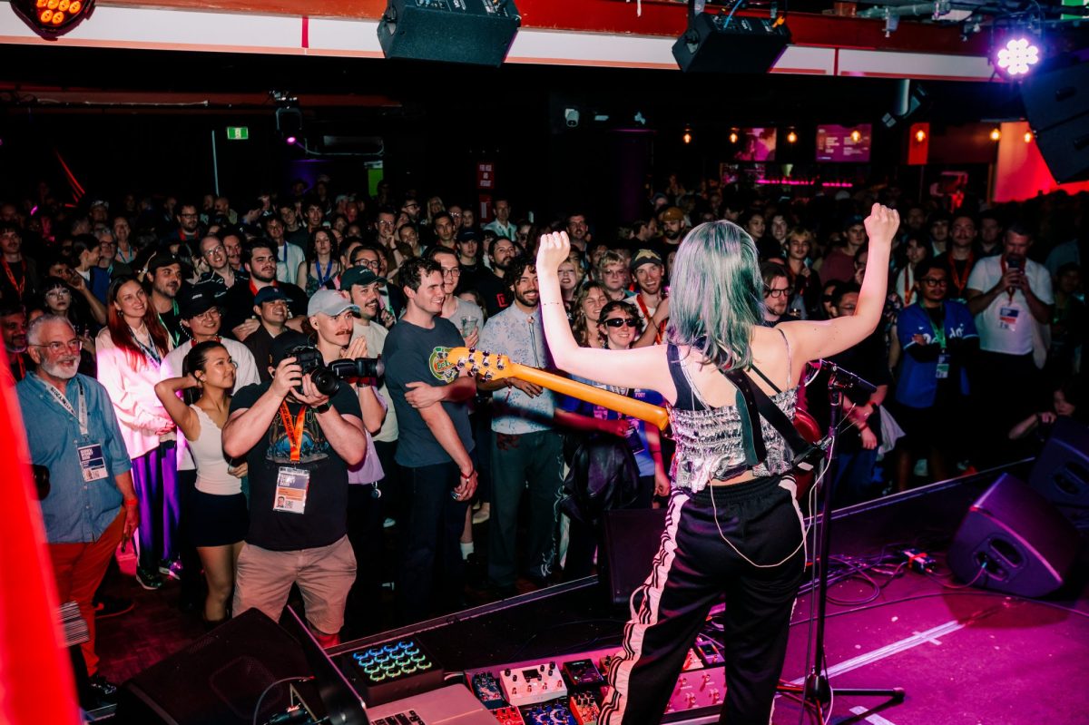 Suichu Spica performs at SXSW Sydney Music Festival 2024 Opening Night Party. A guitarist with green hair stands with their back to the camera, their instrument slung in front of them and hands raised. A large crowd faces them.