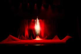 When debate around artists’ rights get heated, we can lose sight of how cultural safety practices act as a positive force to help resolve these situations. The photograph shows seven figures arrayed around a stage, each helping to lift an expansive sheet of red fabric that otherwise covers the stage; red curtains hang behind them, and a spotlight beams down on the central figure. cultural safety