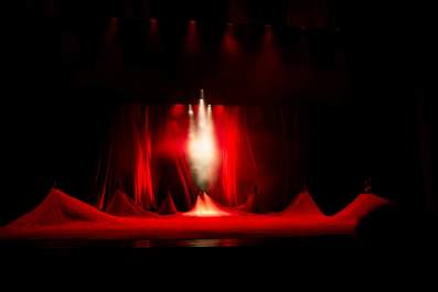 When debate around artists’ rights get heated, we can lose sight of how cultural safety practices act as a positive force to help resolve these situations. The photograph shows seven figures arrayed around a stage, each helping to lift an expansive sheet of red fabric that otherwise covers the stage; red curtains hang behind them, and a spotlight beams down on the central figure. cultural safety