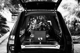A black and white photo of a coffin heaped with flowers in the back of a hearse. Adelaide Writers Week