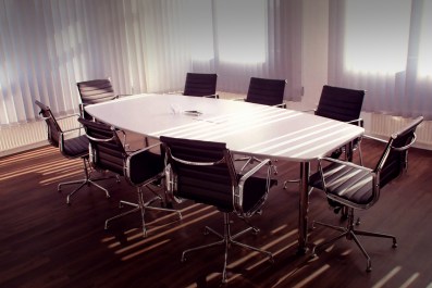 arts board in crisis: a photo of a room with an empty board room table. It's a white oblong table with 8 executive style black chairs around it.