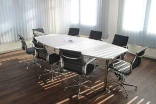 arts board in crisis: a photo of a room with an empty board room table. It's a white oblong table with 8 executive style black chairs around it.