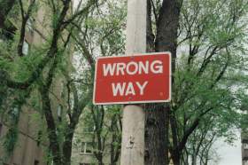 arts governance: photo of a red and white street sign that says "wrong way" against a background of trees in the street.