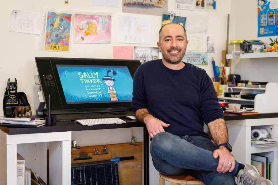 Barking Gecko Arts' ambassador James Foley. A fair-skinned man with short-cropped hair and a high forehead, Foley sits at his desk surrounded by the tools of his trade. A screen sits prominently behind him, displaying one of his characters, Sally Tinker.
