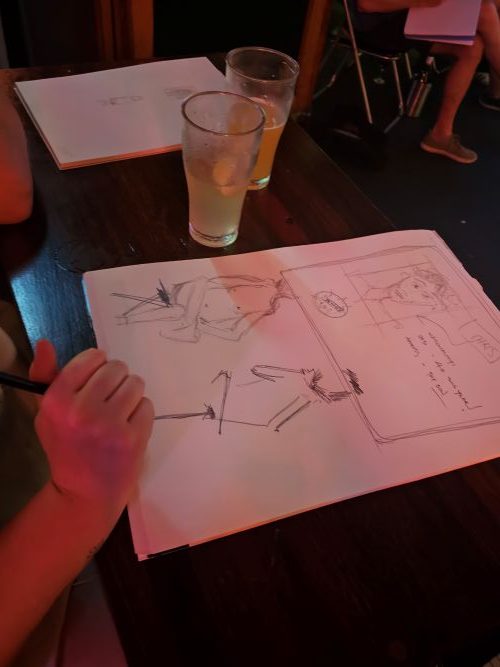 An audience member's work at Life Drawing with a Comedian. The photo shows a large sheet of paper with a series of life drawing sketches, including one underway. The artist's hand is visible, as is the beer they're drinking.