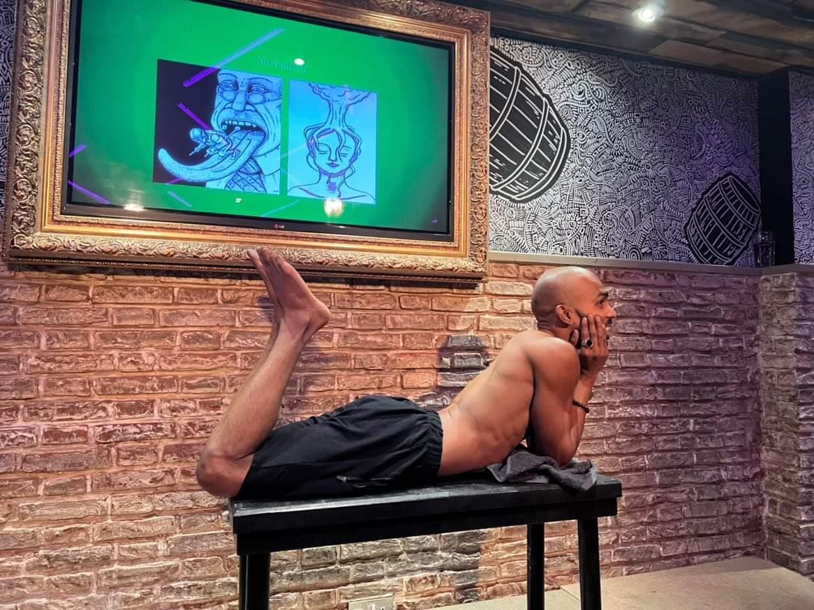 Life Drawing with a Comedian. A shirtless comedian, wearing only black basketball shorts, lies on their stomach on a table, lower legs and feet raised and resting their head on their hands, with their elbows also on the table.