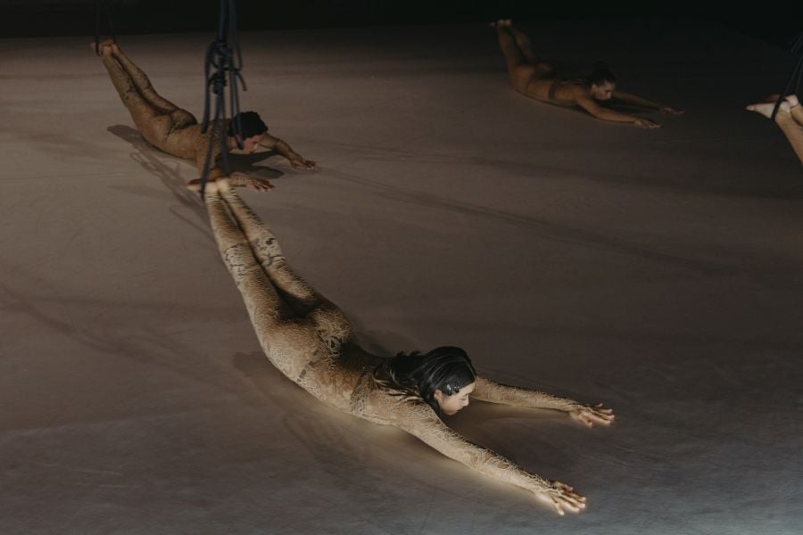 A woman  with shoulder length black hair and wearing a lace-patterned body stocking lies on stage on her stomach; her arms are extended and her feet are suspended and raised off the stage in a loop of rope. Other dancers in similar positions are visible behind and around her. Yilin Kong and Zachary Lopez in ADT's Faraway by Jenni Large at Adelaide Festival 2026.
