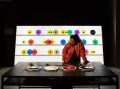 Carsten Nicolai's installation Bausatz Noto (1998) at The Vinyl Factory: Reverb. A dark skinned woman wearing a red leather jacket and headphones listens to music at a desk of turntables. A rack of brightly coloured vinyl records stands behind her.