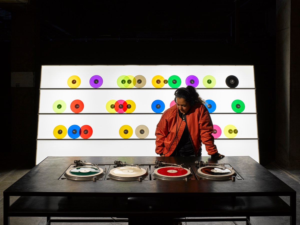 Carsten Nicolai's installation Bausatz Noto (1998) at The Vinyl Factory: Reverb. A dark skinned woman wearing a red leather jacket and headphones listens to music at a desk of turntables. A rack of brightly coloured vinyl records stands behind her.