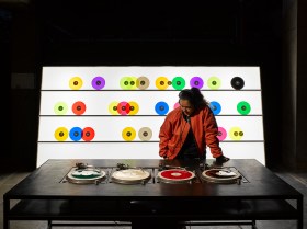 Carsten Nicolai's installation Bausatz Noto (1998) at The Vinyl Factory: Reverb. A dark skinned woman wearing a red leather jacket and headphones listens to music at a desk of turntables. A rack of brightly coloured vinyl records stands behind her.