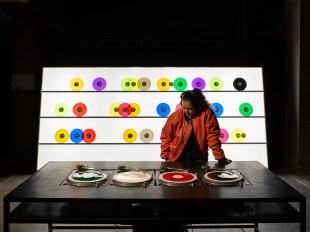 Carsten Nicolai's installation Bausatz Noto (1998) at The Vinyl Factory: Reverb. A dark skinned woman wearing a red leather jacket and headphones listens to music at a desk of turntables. A rack of brightly coloured vinyl records stands behind her.