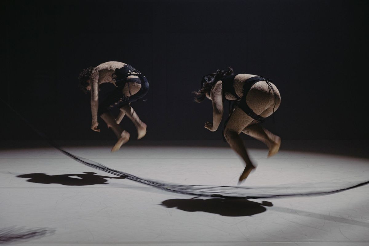 Two dancers clad in fetish wear-inspired costumes crouch forward as they jump a woollen rope inside a circle of light on stage. Joshua Doctor and Yilin Kong in ADT's Faraway by Jenni Large at Adelaide Festival 2026.