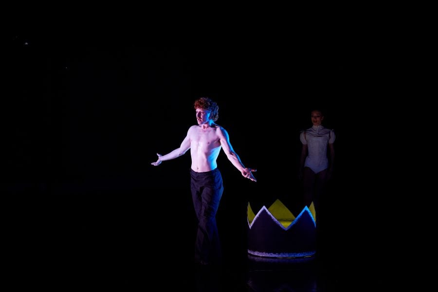 Adam Alzaim and Asja Petrovski in Polly Hilton’s Paper Moon for West Australian Ballet's Ballet at the Quarry: Incandescence, 2026. Alzaim is shirtless and wears dark pants; he has red hair, and stands with his chest out and his arms partially raised. Behind him, a giant crown, partially illiminated from within, sits on the stage; Petrovski stands behind it in shadow, wearing white.