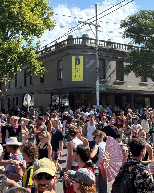 Australian Print Workshop during the Midsumma closing event Victoria's Pride in February 2026. A grey Victorian-era building, complete with rooftop balustrades, occupies a crowded street corner. A yellow sign featuring the building's name and Australian Print Workshop's logo hangs horizontally on the building's street front.