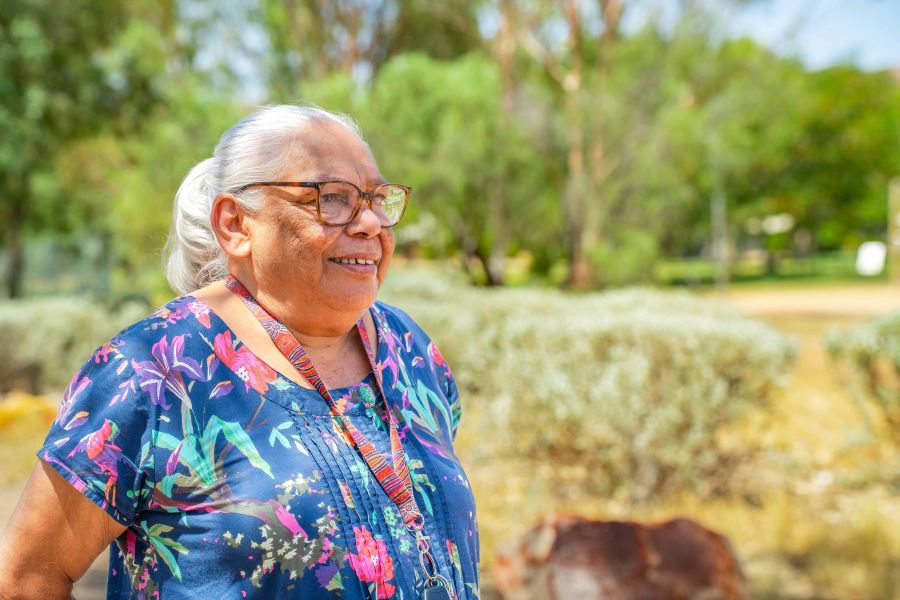 Children's Ground's new Co-Chair Evelyn Schaber. An older Aboriginal woman with white hair tied behind her head. She wears glasses, a striped lanyard around her neck and a colourful, short-sleeved dress printed with floral designs. Evelyn is looking off into the distance and smiling; the landscape behind her is out of focus. arts sector appointments